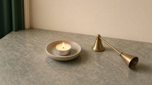 A lit tealight in a ceramic saucer on a clear stone surface with a brass candle snuffer